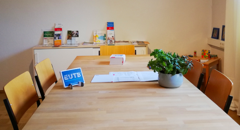 Der Beratungsraum der EUTB. Ein großer Tisch mit Stühlen und einer Pflanze. Im Hintergrund sind verschiedene Flyer sowie eine Kinderspielecke zu sehen.