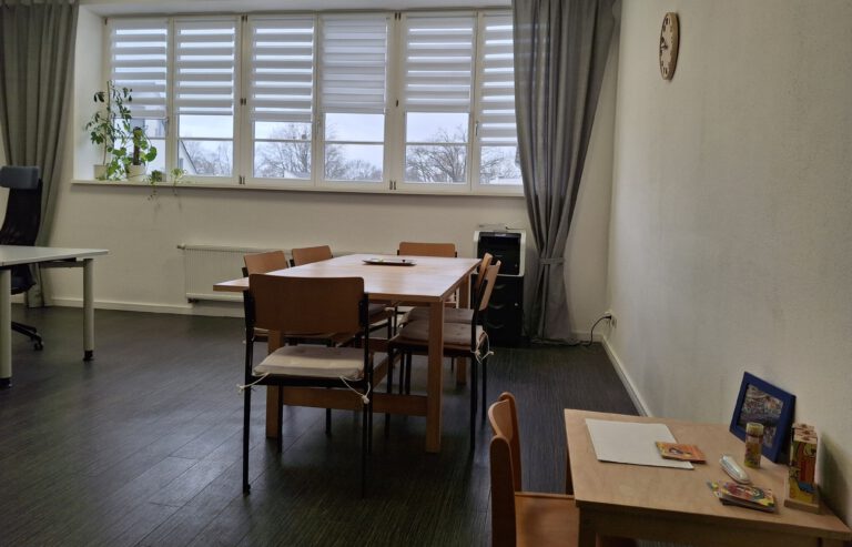 Im Vordergrund steht ein kleiner Tisch mit Spielzeug für Kinder. Im Hintergrund steht ein großer Tisch mit Sitzplätzen vor einer großen Fensterfront.